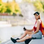 Pretty young hipster asian woman in black hat vape ecig, vaping device at the sunset. Toned image