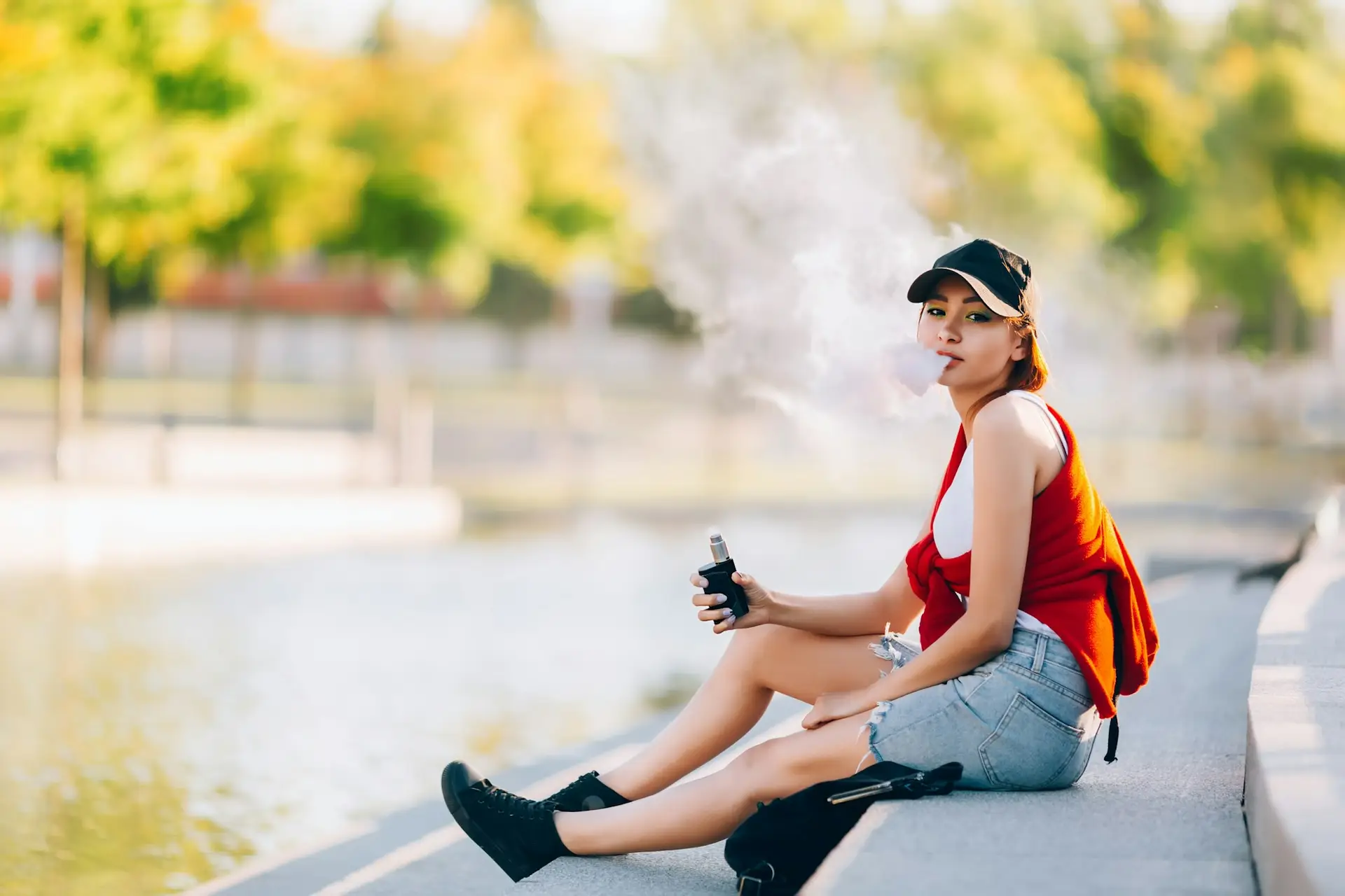 Pretty young hipster asian woman in black hat vape ecig, vaping device at the sunset. Toned image